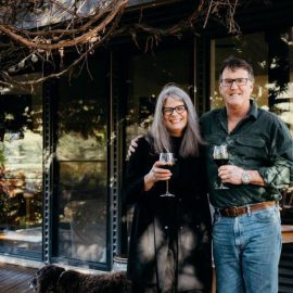 Owners Julie & Dean standing together under verandah with a glass of red wine in hand.